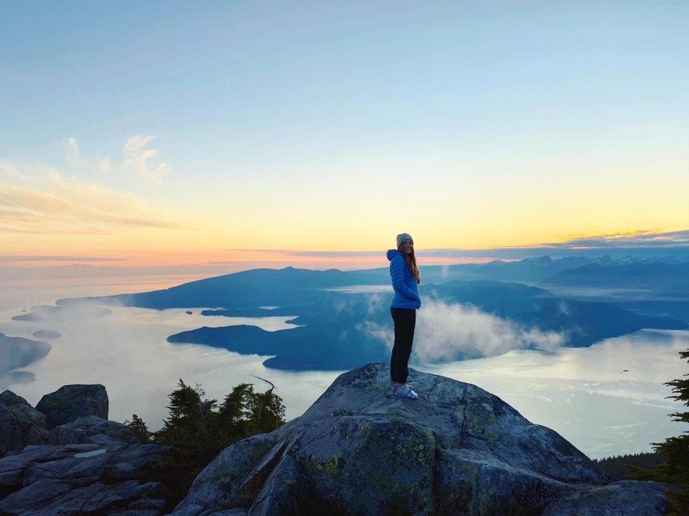 Alicia in a blue jacket and black hiking pants standing on top of a mountain overlooking the water