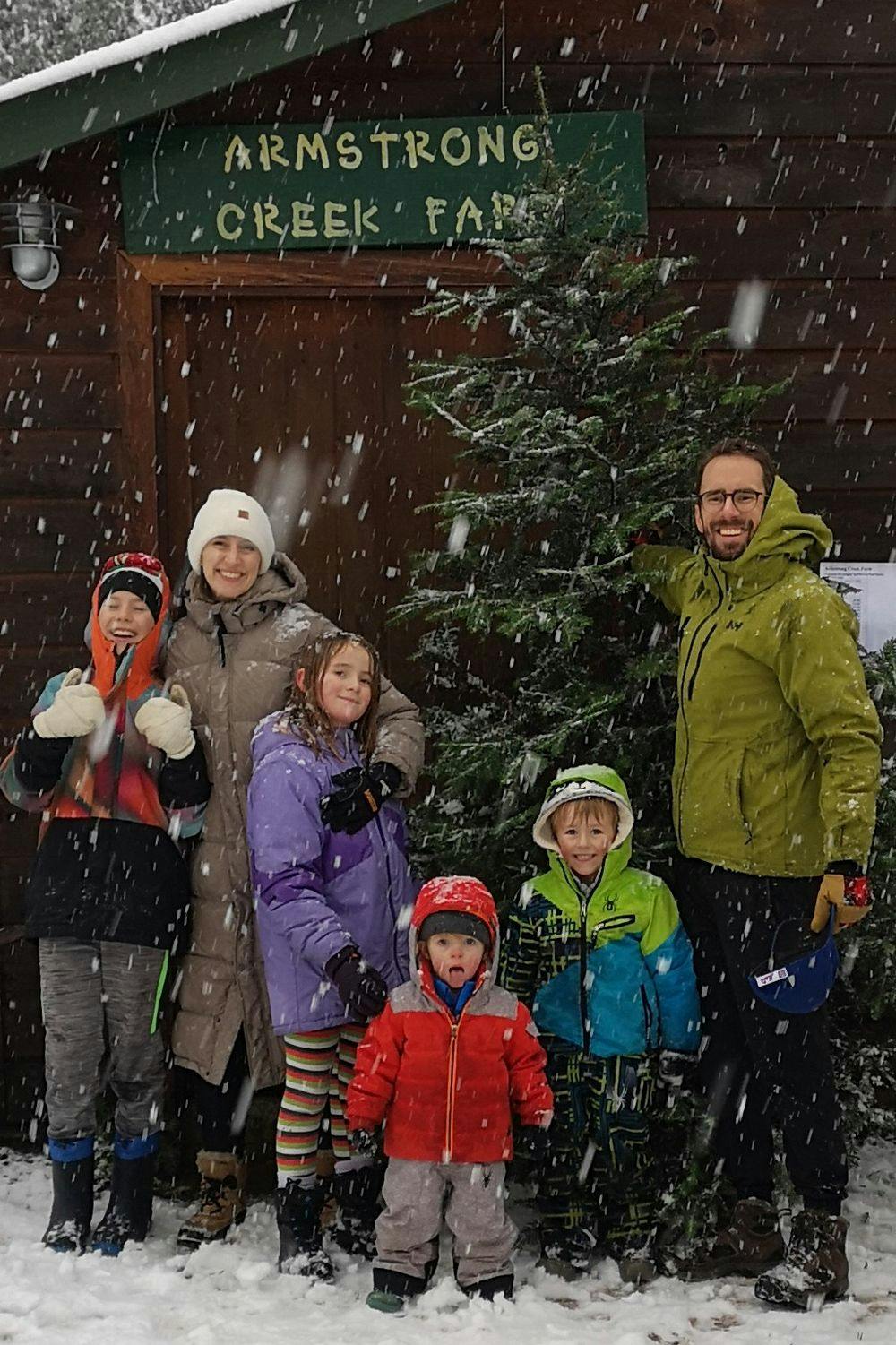 NJ, her husband, and her four children smiling in the snow in front of an evergreen tree