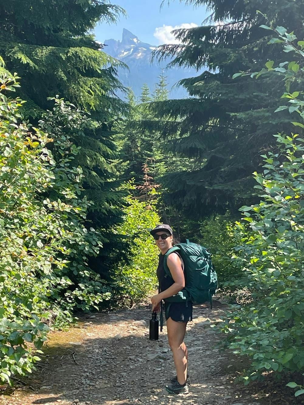 Pietra smiling and wearing a large green backpack on a hiking trail with green trees in the background