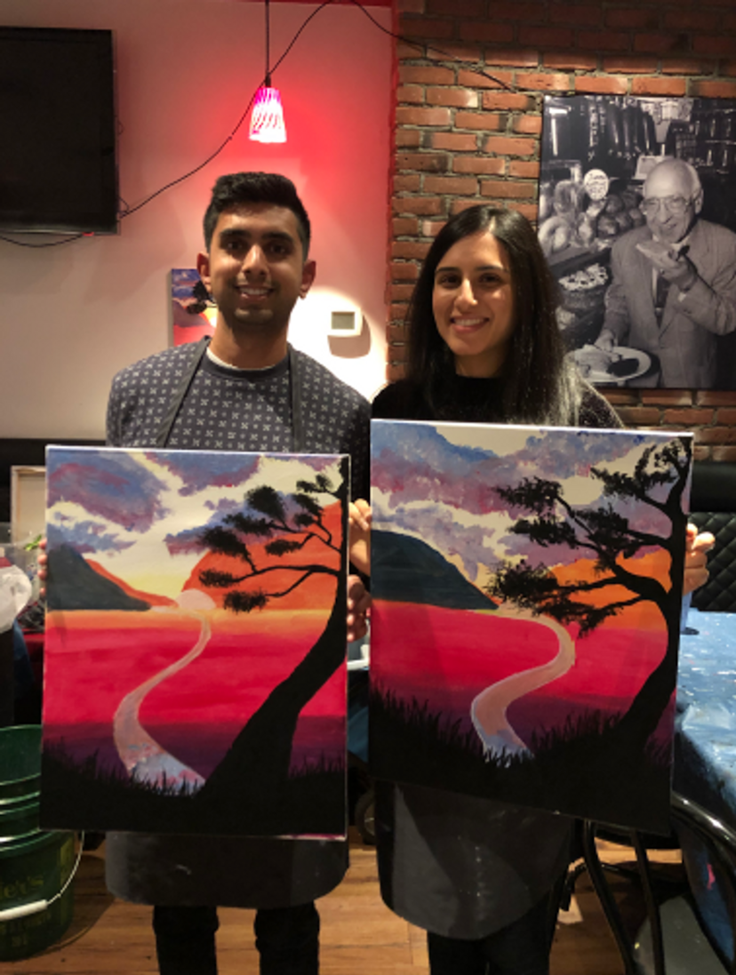 Narissa and her partner holding their paintings of a red field and tree with the sun and clouds