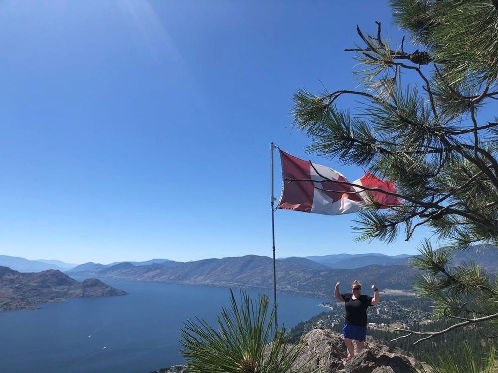 Katie O'Connell standing on top of a mountain with the Canadian flag overlooking a lake and more mountains