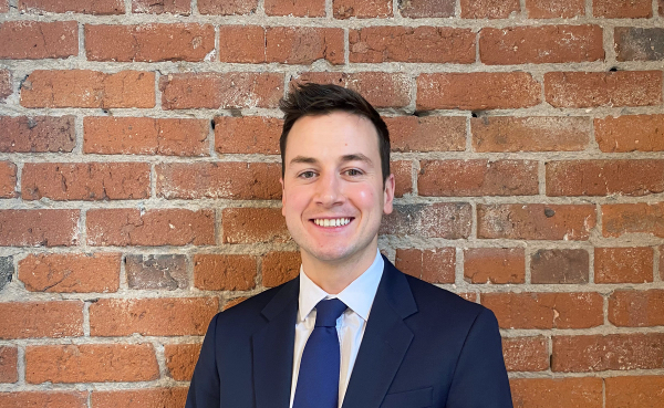 Headshot of Kieran smiling in a suit in front of a brick wall