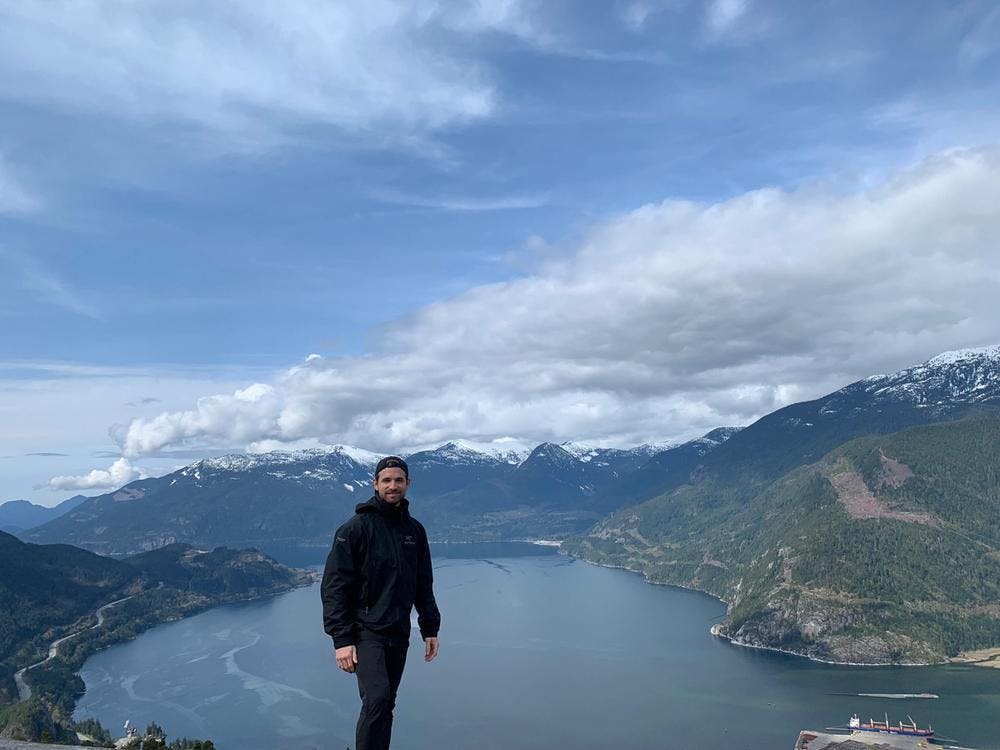Marc standing on top of a mountain with a view of a lake and other mountains
