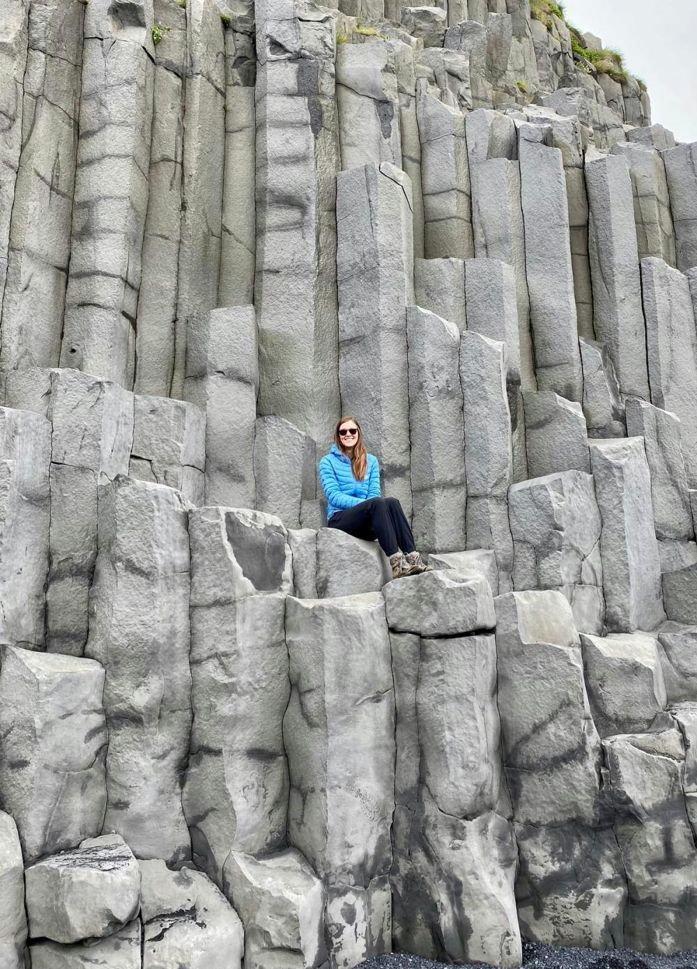 Alicia Stirling in a blue jacket and black pants sitting on gray basalt columns in Iceland
