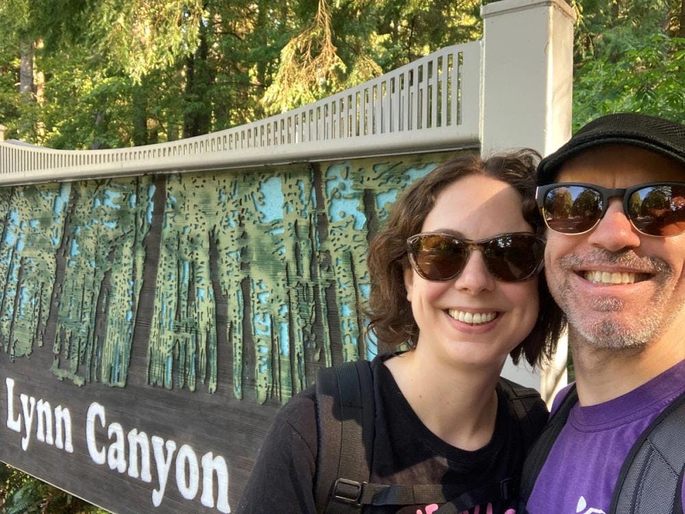 Jennifer and partner wearing sunglasses and smiling in front of the sign for Lynn Canyon