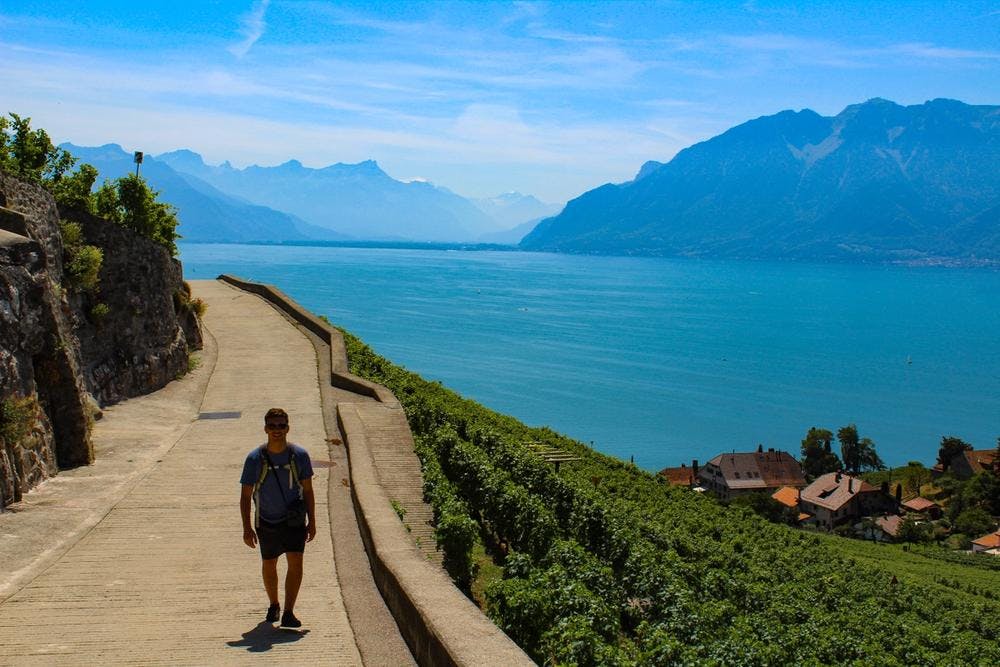 Liam in hiking gear on a walkway overlooking bright blue water of Lake Geneva in Switzerland and mountains in the background