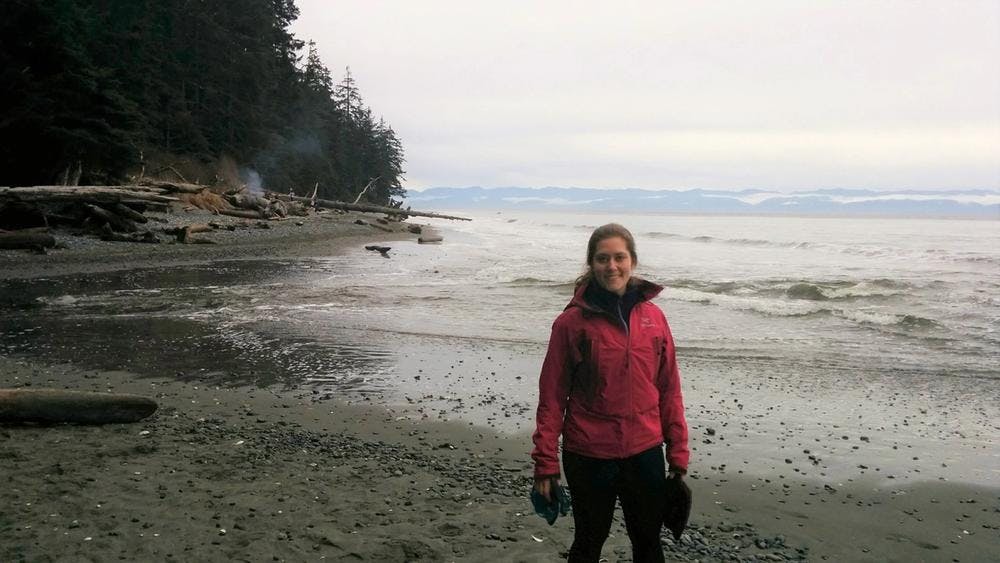Janel standing on a beach with the forest and water behind her